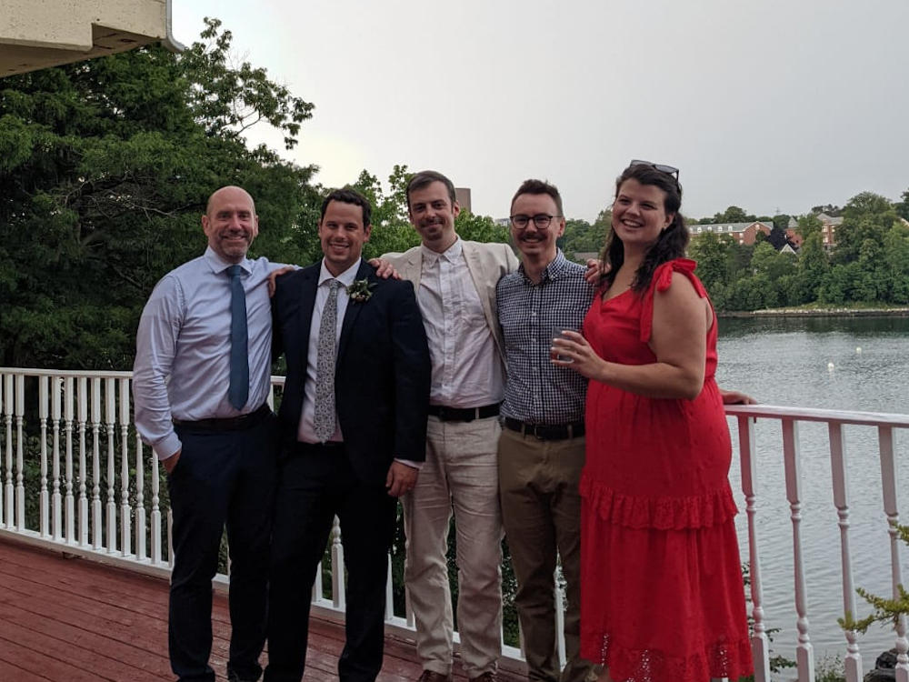 Five extremely handsome people in nice clothes, posing for a picture in front of a body of water